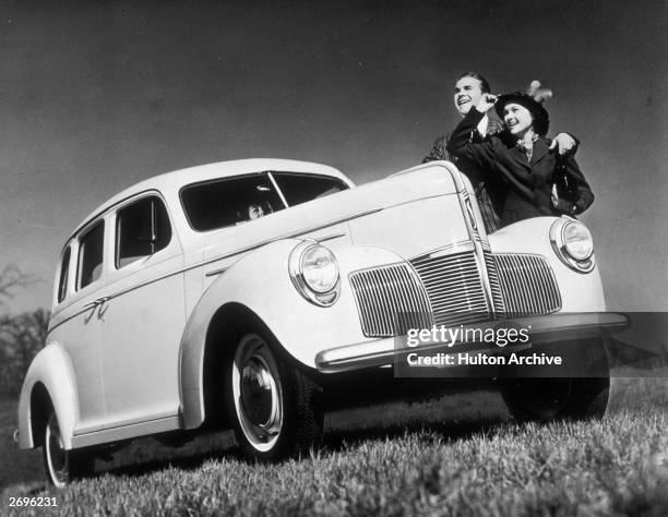 Low-angle view of a happy couple smiling behind a new 1940 Studebaker outdoors in a field. Someone sits behind the wheel of the car.