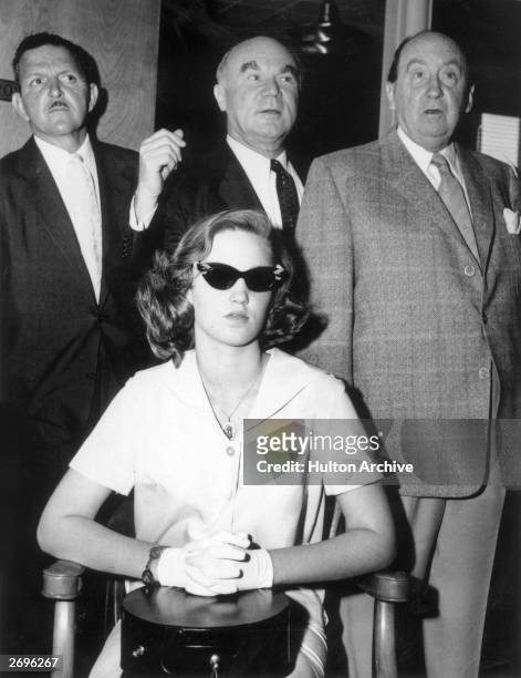 Cheryl Crane, daughter of actor Lana Turner, sits in a chair with her hands folded in her lap, wearing dark sunglasses, while three unidentified men...