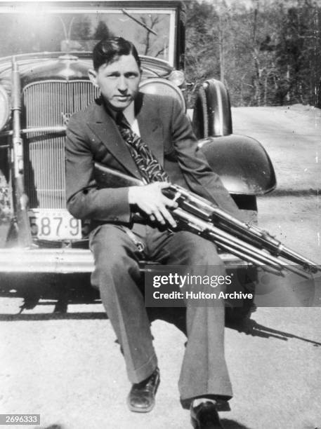 Portrait of American criminal Clyde Barrow holding a machine gun while sitting on the front fender of a car.