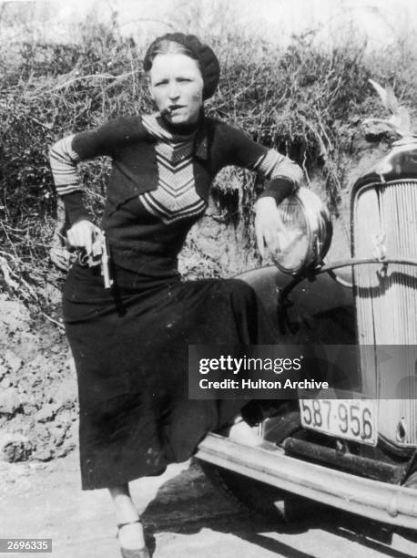 Full-length portrait of American criminal Bonnie Parker smoking a cigar while leaning on the front fender of a car and holding a pistol.