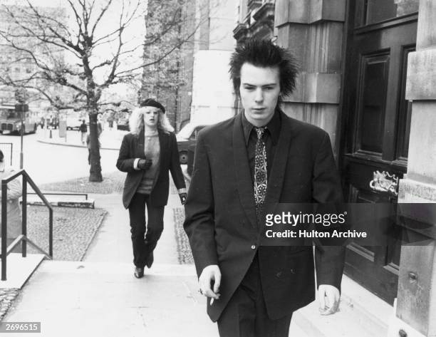 British musician Sid Vicious, bass player and vocalist for the British punk rock band The Sex Pistols, smokes a cigarette while walking up a flight...