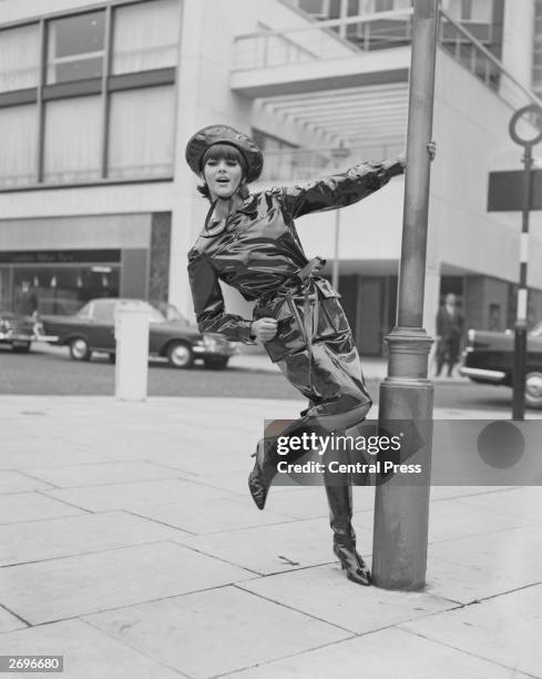 Model Jackie Bowyer swings on a lamppost wearing a black oilskin wet-weather outfit from Mary Quant, of matching coat and hat with shiny, high-heeled...