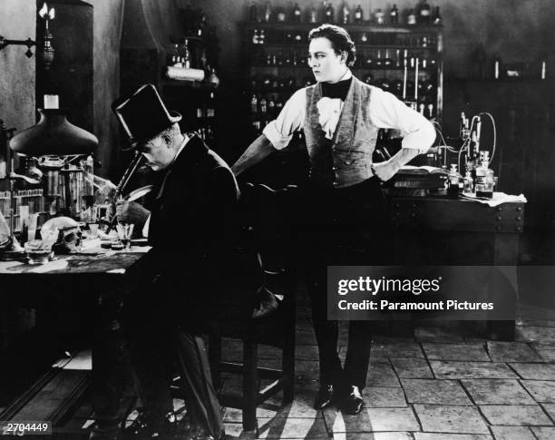 American actor John Barrymore stands in the middle of a laboratory as an unidentified actor in a top hat looks through a microscope in a still from...