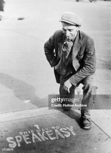 Man kneeling on the pavement, next to a sign showing the way to a speakeasy, during the Prohibition in America.