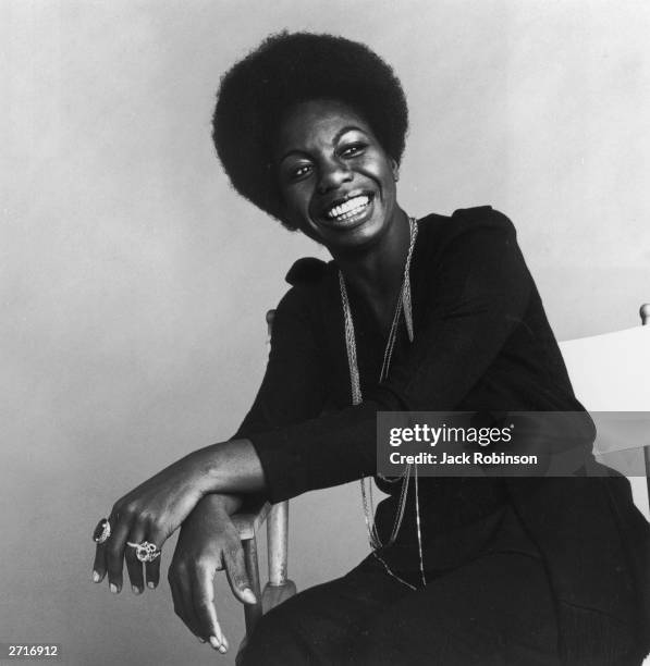 Portrait of American vocalist Nina Simone sitting in a white director's chair and smiling.