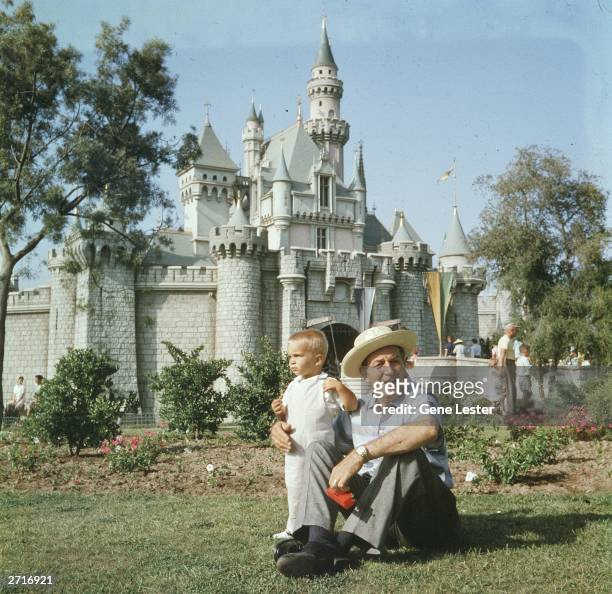 American animator and film studio founder Walt Disney sits on a grassy lawn with his grandson, in front of the Magic Kingdom's castle at Disneyland,...