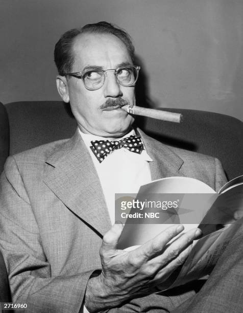 Portrait of American comedian Groucho Marx, wearing eyeglasses, business suit and polka dot bow tie, seated in an armchair and smoking a cigar, looks...