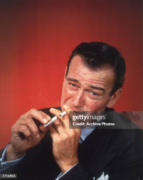 Headshot studio portrait of American actor John Wayne in front of a red background, dressed in a sport coat, lighting a cigarette with an already lit...