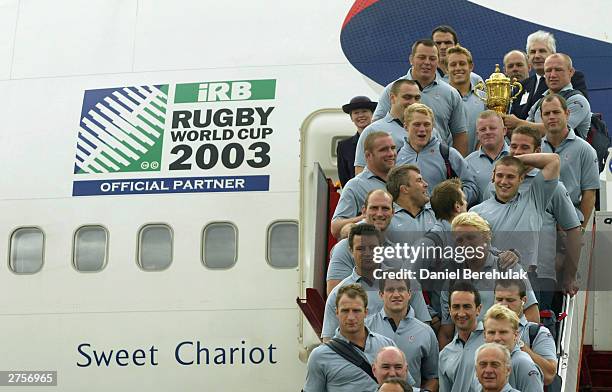 The England team hold aloft the Webb Ellis Trophy on the stairs of the British Airways BA16 aircraft during the departure of the England Rugby team...