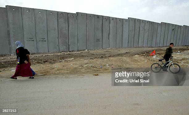 Palestinians pass by the Israeli separation barrier December 22, 2003 between their West Bank village of Nazlat Issa and the neighboring Israeli Arab...
