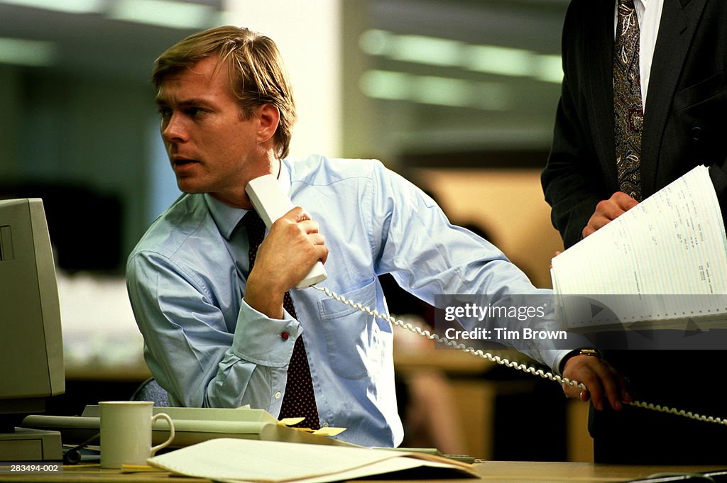 Two men discussing printout,one at desk holding phone