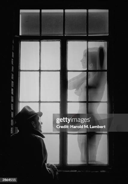 Man wearing a hat and coat stands outside a window at night and watches a woman undress, circa 1960s.