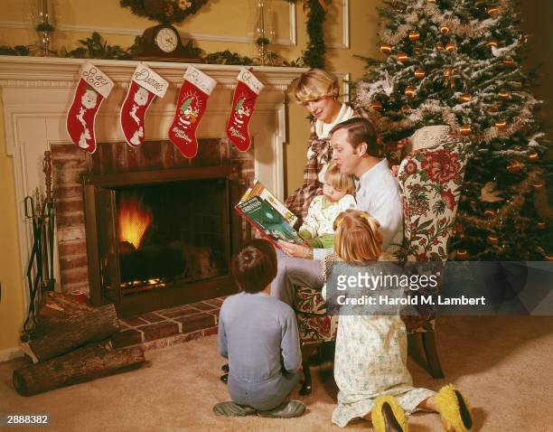 Father sits in an armchair by the fireplace, reading a storybook to his children, circa 1970. There is a decorated Christmas tree in their living...