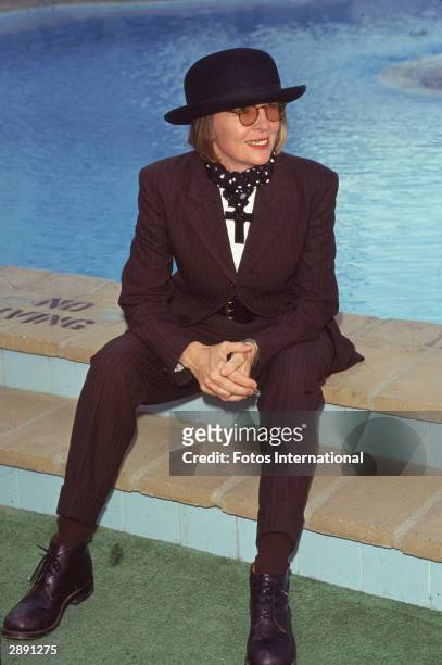 Portrait of American actor and director Diane Keaton as she sits near a swimming pool, 1996. She wears a purple & black striped suit, bowler hat,...