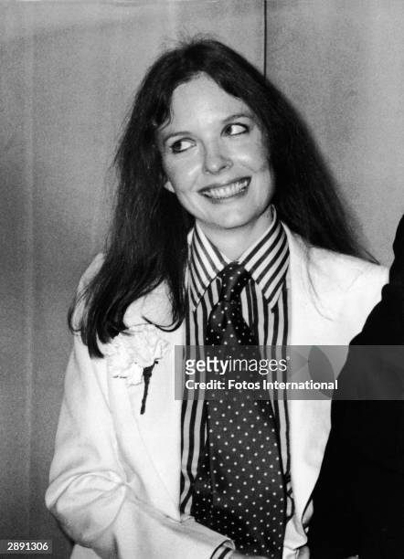 American actor and director Diane Keaton poses with a big smile and tilted head at the 48th Academy Awards and wears a white suit with a carnation in...