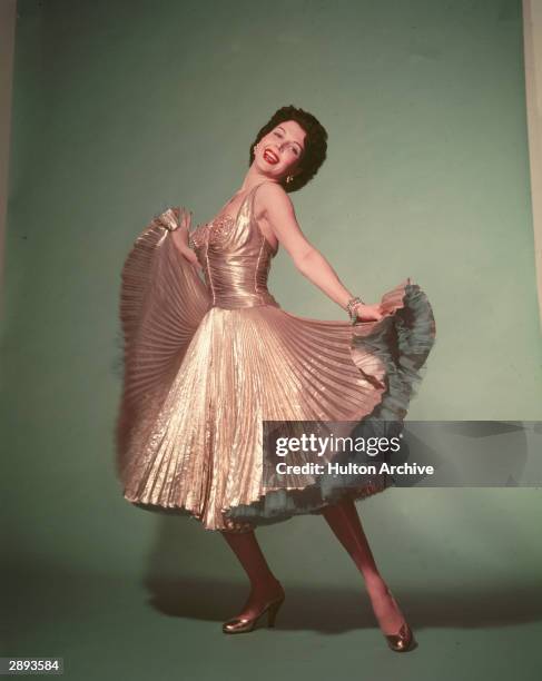 Portrait of American actor and dancer Ann Miller , circa 1940s. She wears a pleated gold dress and petticoat and lifts the hem of her dress with her...