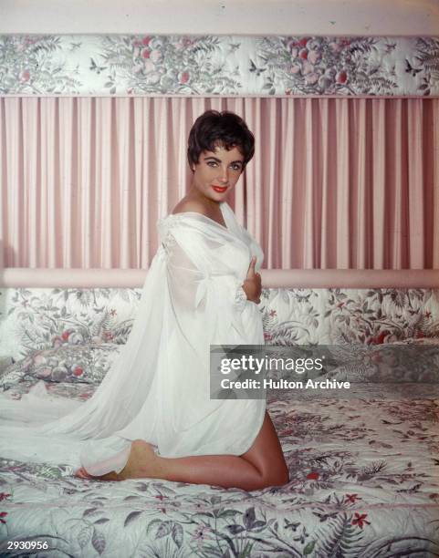 British-born actor Elizabeth Taylor kneels on a bed and wears a sheer white nightgown and robe, circa 1950s.