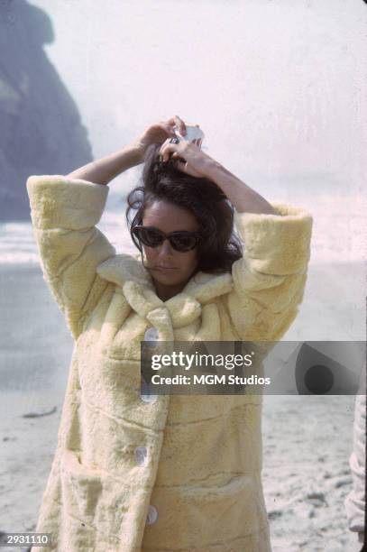 British-born actor Elizabeth Taylor, in a furry yellow coat with oversized buttons, adjusts her hair while on the set of 'The Sandpiper,' directed...