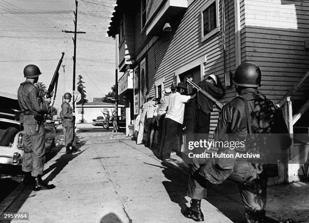 Armed National Guardsmen force a line of Black men to stand against the wall of a building during the Watts race riots, Los Angeles, California,...