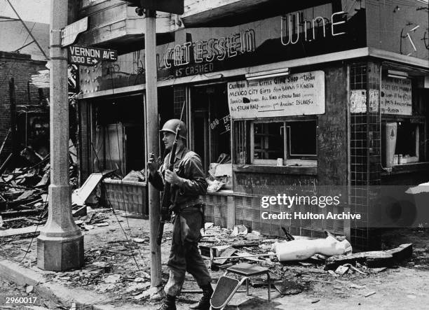 An armed National Guard patrolman leans against a street sign, smoking a cigarette and standing in rubble following the Watts riots, Los Angeles,...