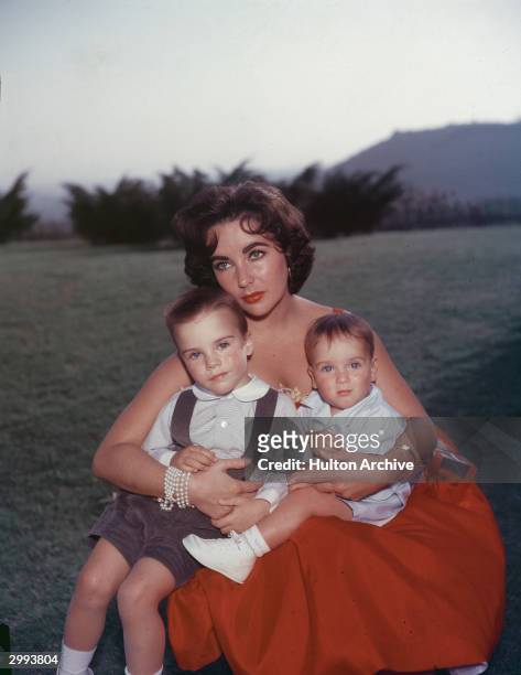 British-born actor Elizabeth Taylor sits in a red evening dress with her sons Michael and Christopher Wilding on her lap, mid 1950's.