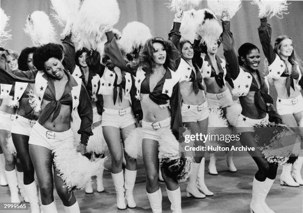 The Dallas Cowboy Cheerleaders perform on stage in matching costumes, cheering and shaking pom-poms, 1970s.