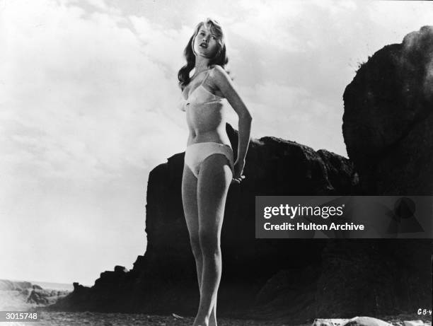 French-born actor Brigitte Bardot wears a white bikini and stands on a rocky beach in a still from the film, 'The Girl in the Bikini', directed by...