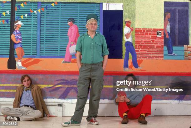 British-born pop artist David Hockney stands in front of a wall-sized painting while two unidentified men sit on either side of him and lean against...