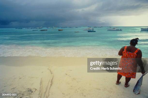 west indies,barbados,woman on beach holding fish,boats moored in bay - fish barbados stock pictures, royalty-free photos & images