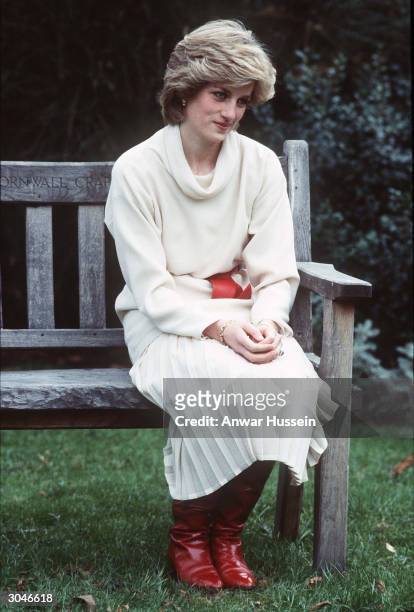 Diana, Princess of Wales, wearing a cream cowl neck jumper and pleated skirt with a red belt and boots, sits on a wooden bench that was a wedding...
