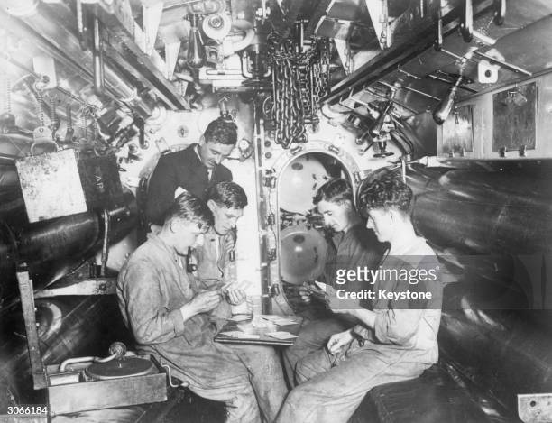 Crew of a submarine docked in Portsmouth having a game of cards while listening to a gramophone record in the torpedo firing room.