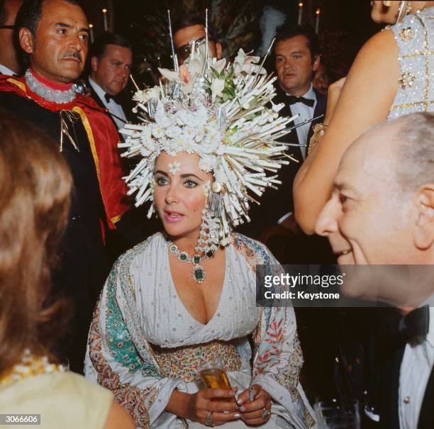 English movie star Elizabeth Taylor attends a social function wearing an elaborate headdress of pearls and fake flowers, a jewelled dress and an...