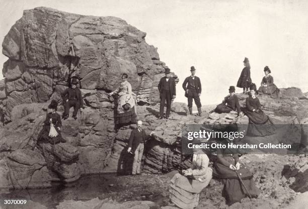 Fully-dressed Victorian holidaymakers sunbathing on a rocky shore.