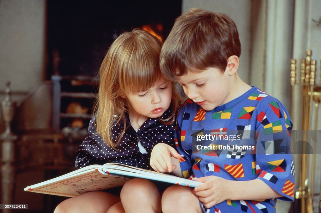 Boy(4-5)and girl(2-3)sat by fire in pyjamas reading book together