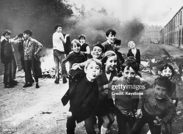 Group of children playing up to the camera while a fire smoulders in the street behind them, Belfast, Northern Ireland, December 1971.