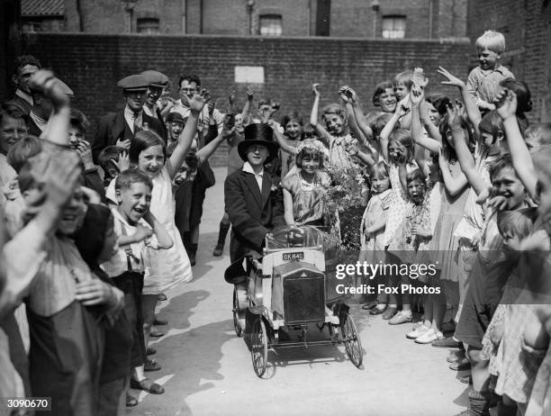 Children in the slum streets of Caledonian Road, London play 'getting married'. Here the bridal pair drive away in their car to the cheers of friends.