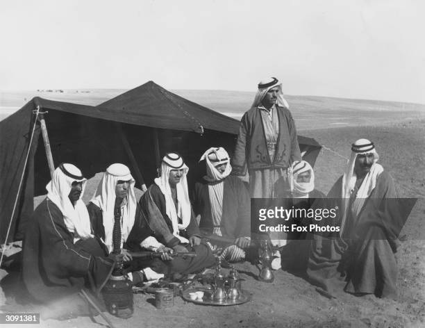 Group of Bedouin men in traditional headdresses seated around coffee pots in front of a tent.