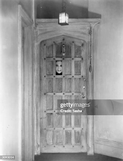 Composer and actor Ivor Novello with just his face seen peering through a spy hole in a door.