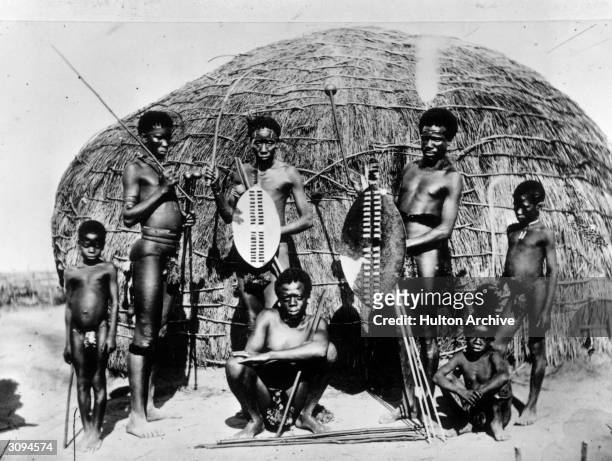 Group of Zulus with spears and shields outside one of their 'beehive' houses. These are made of thatch laid over a wooden framework and are usually...
