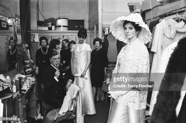 Backstage at a fashion show, Nola models a Norman Hartnell evening dress with a lace bodice and frilly hat. Hartnell himself keeps an eye on...