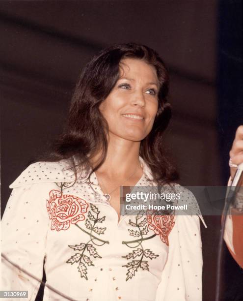 American country music singer and guitarist Loretta Lynn smiles as she looks at an unidentified person off-camera who holds a microphone, late 1970s.