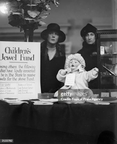 Children's Jewel Fund. Lady Diana Manners and Lady Violet Bonham Carter. Stand behind a counter on which sits a doll round whose neck is a sign which...
