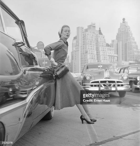 Vogue fashion model wearing a day suit leans against a Buick motor car. A Pontiac is parked in the background.