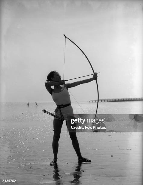 Woman in a bathing suit takes aim with her bow and arrow on the beach at Worthing, Sussex.