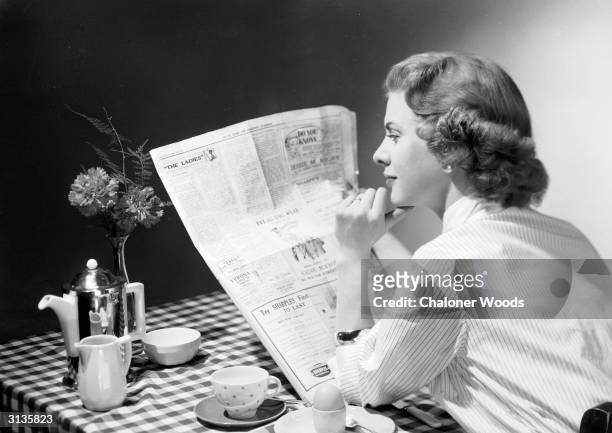 Woman wearing a striped shirt blouse reads a newspaper over the breakfast table before tackling her boiled egg and coffee.