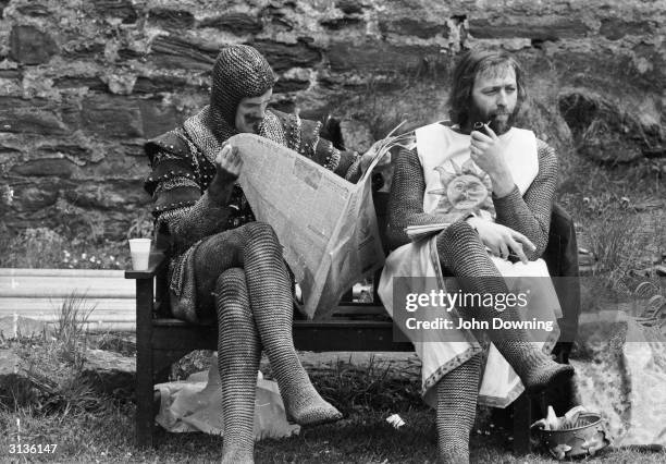 Chainmail-clad John Cleese reads a newspaper while Graham Chapman smokes a quiet pipe on the set of 'Monty Python and the Holy Grail'.
