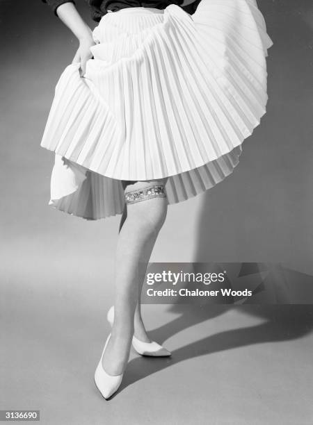 Sixties fashion model lifting her pleated hemline to reveal an ornate garter above the knee.