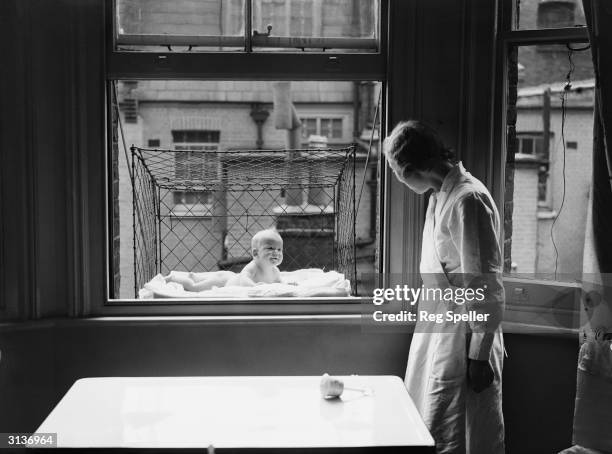 Nanny supervising a baby suspended in a wire cage attached to the outside of a high tenement block window. The cages are distributed to members of...