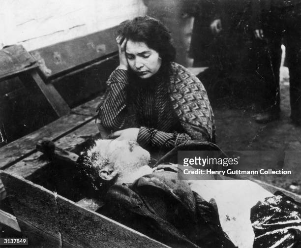 Woman weeps over the body of a man lying on a wooden bench, the victim of a pogrom in Eastern europe.