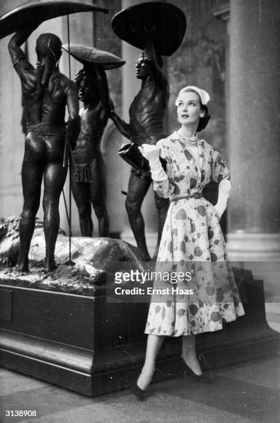 Vogue fashion model wearing a flower patterned dress with gloves and a handbag, standing next to a sculpture in a New York museum.